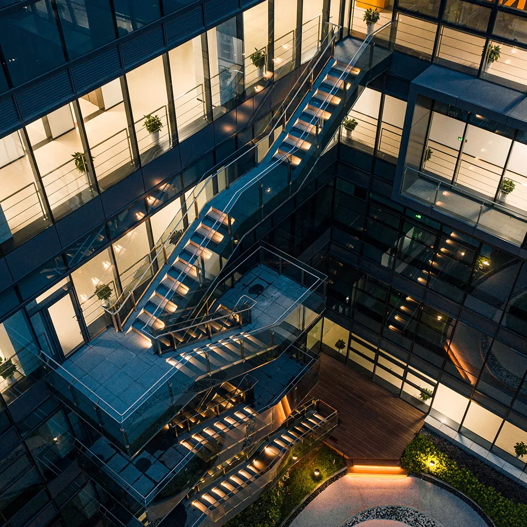 Modernes Bürogebäude mit Glasfassaden, beleuchteten Treppen und mehreren Etagen. Die Architektur symbolisiert die vernetzten Strukturen einer Full-Service-Marketing-Agentur.