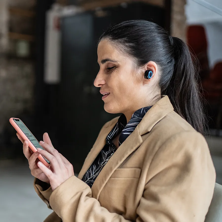 Frau mit dunklen Haaren im Pferdeschwanz, trägt einen beigen Blazer und kabellose Ohrhörer, sitzt an einem Tisch und benutzt ein Smartphone. Sie ist sehbeeinträchtigt. Im Hintergrund sitzt eine weitere Person.